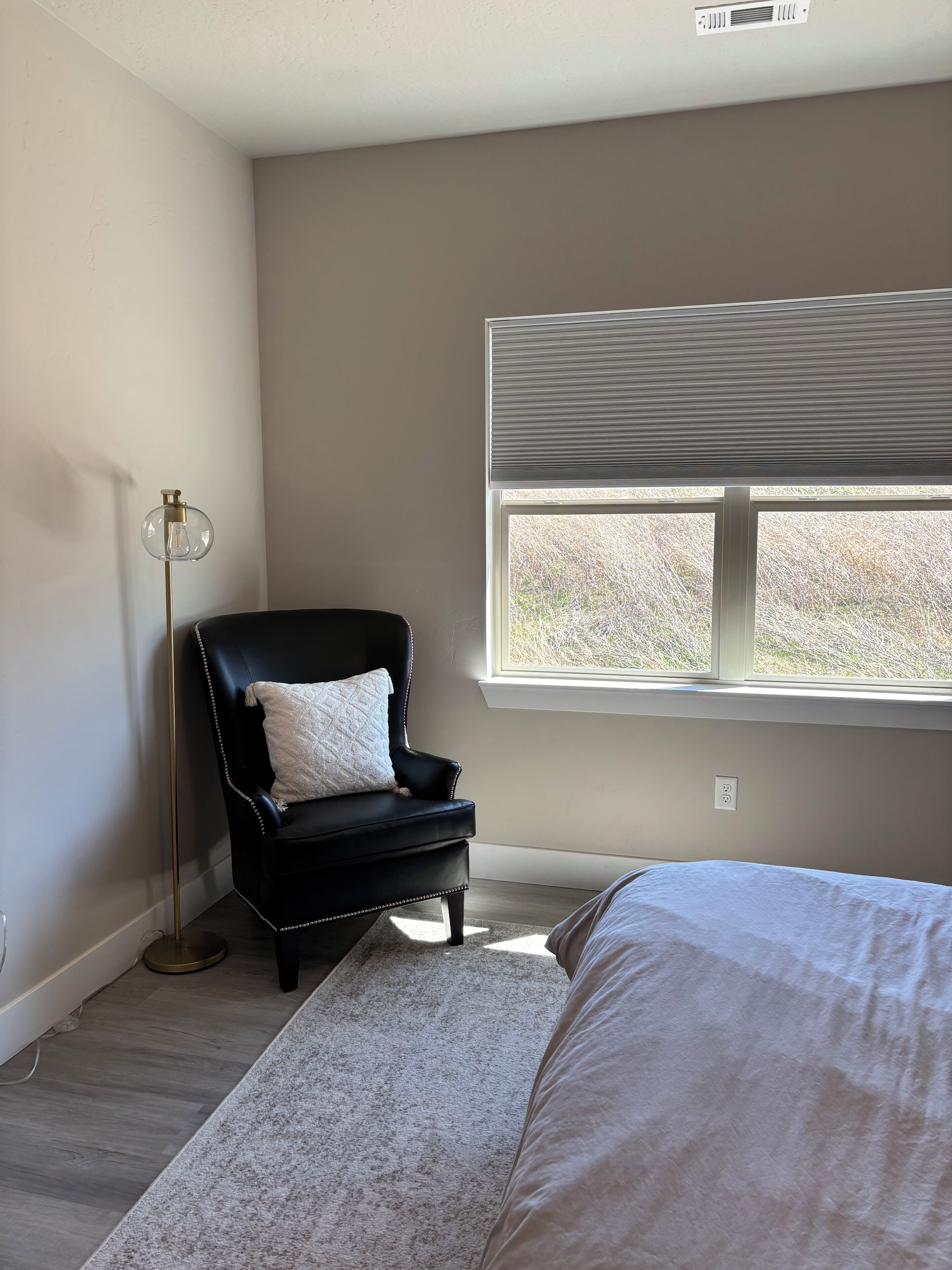 A bedroom with a bed, a black leather chair, a floor lamp, and a window with blinds.