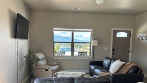 A living room with a gray sectional sofa, a white recliner, and a black leather couch. A flat-screen TV is mounted on the wall, and a window offers a view of the outdoors. - Charming 2-bedroom house in Stevensville with AC, WiFi (Stevensville)