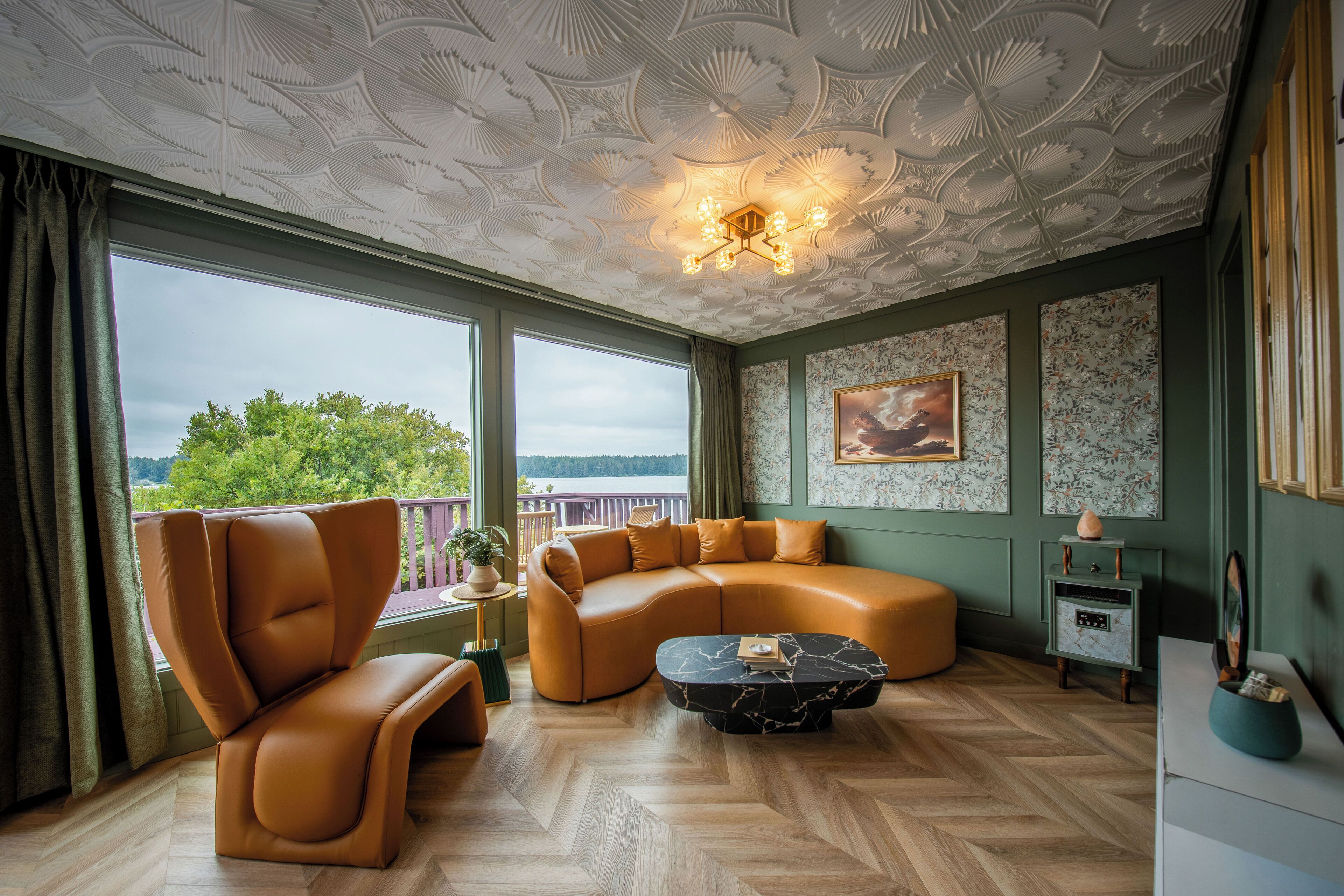 A spacious living room with a large sectional sofa, a curved coffee table, and a modern chair. The room features a decorative ceiling and large windows offering a view of greenery and a body of water.