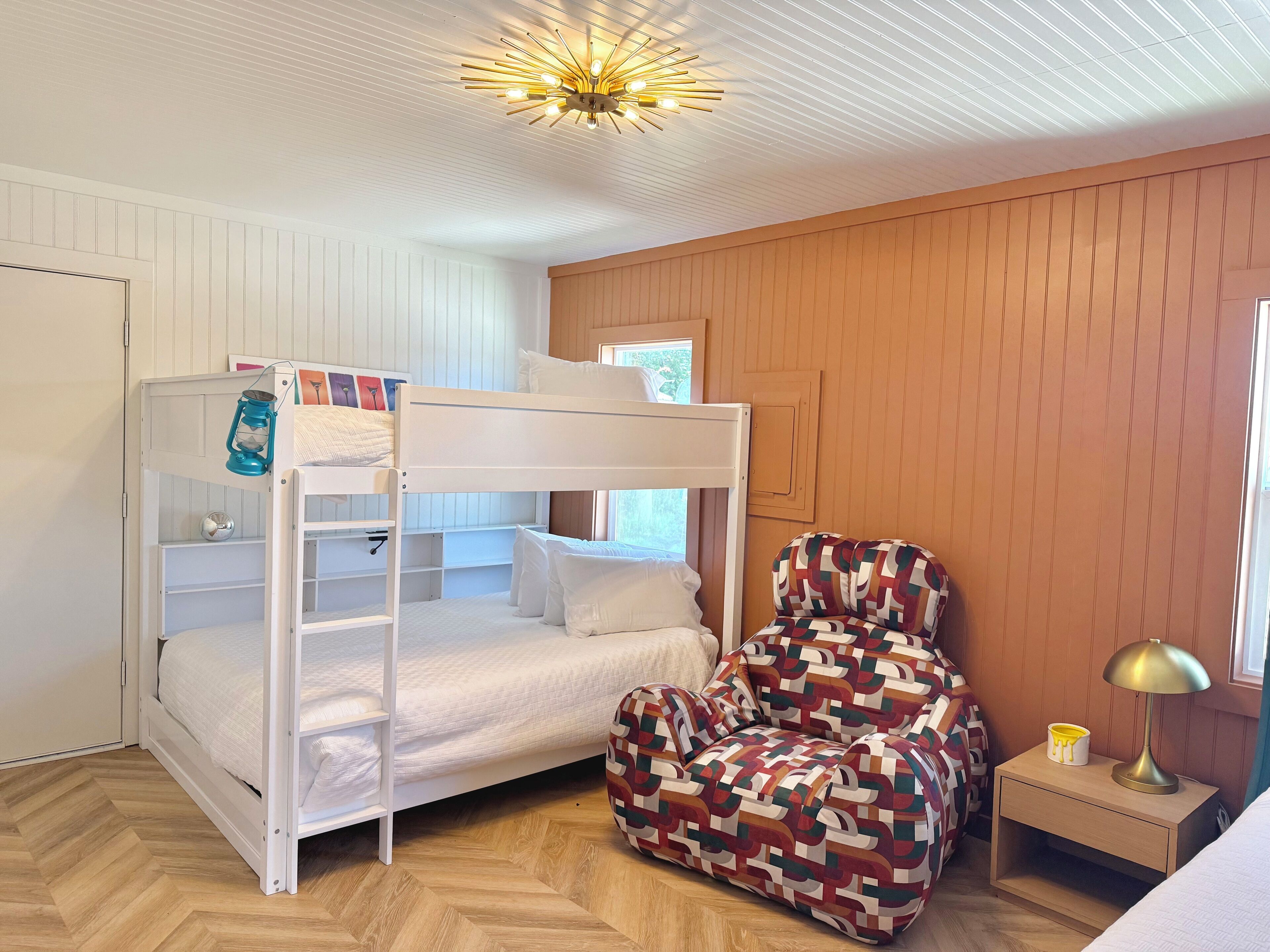 A bunk bed with a lantern, a patterned armchair, a bedside table with a lamp, and a window with a view.
