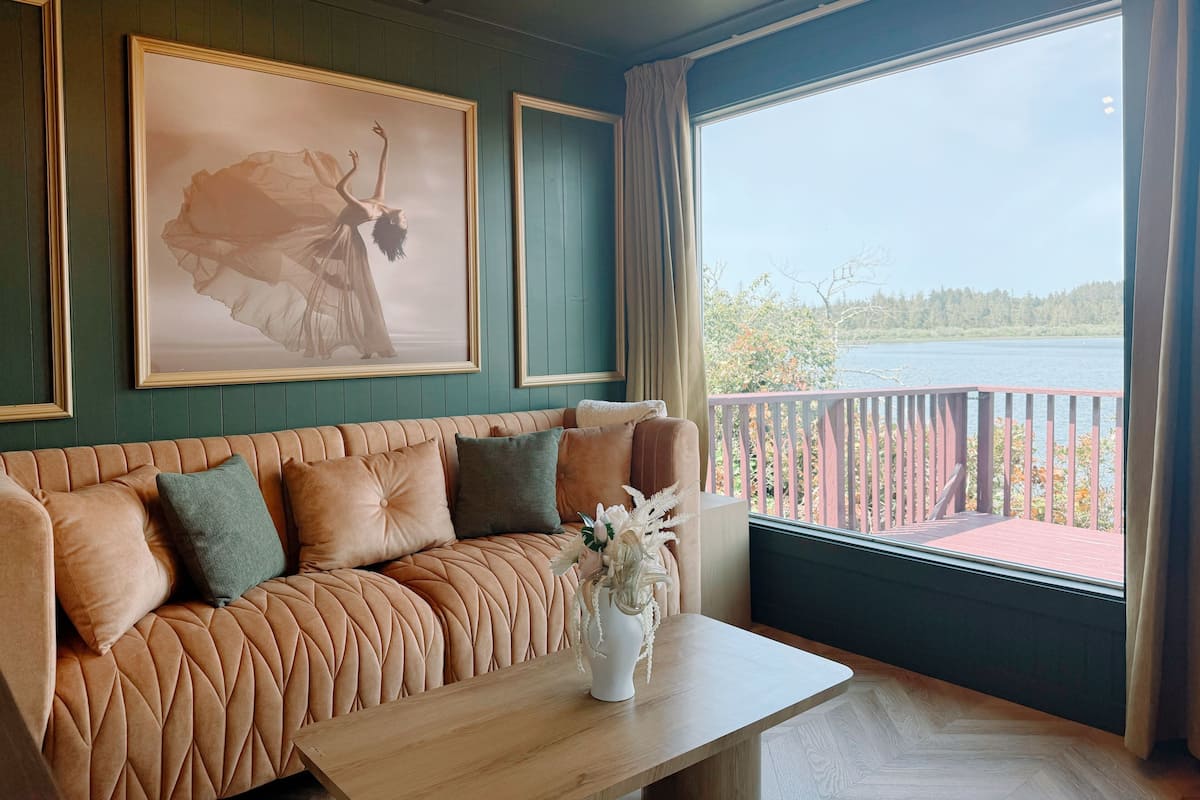 A room with a tufted sofa, a wooden table, and a large window with a view of a lake.
