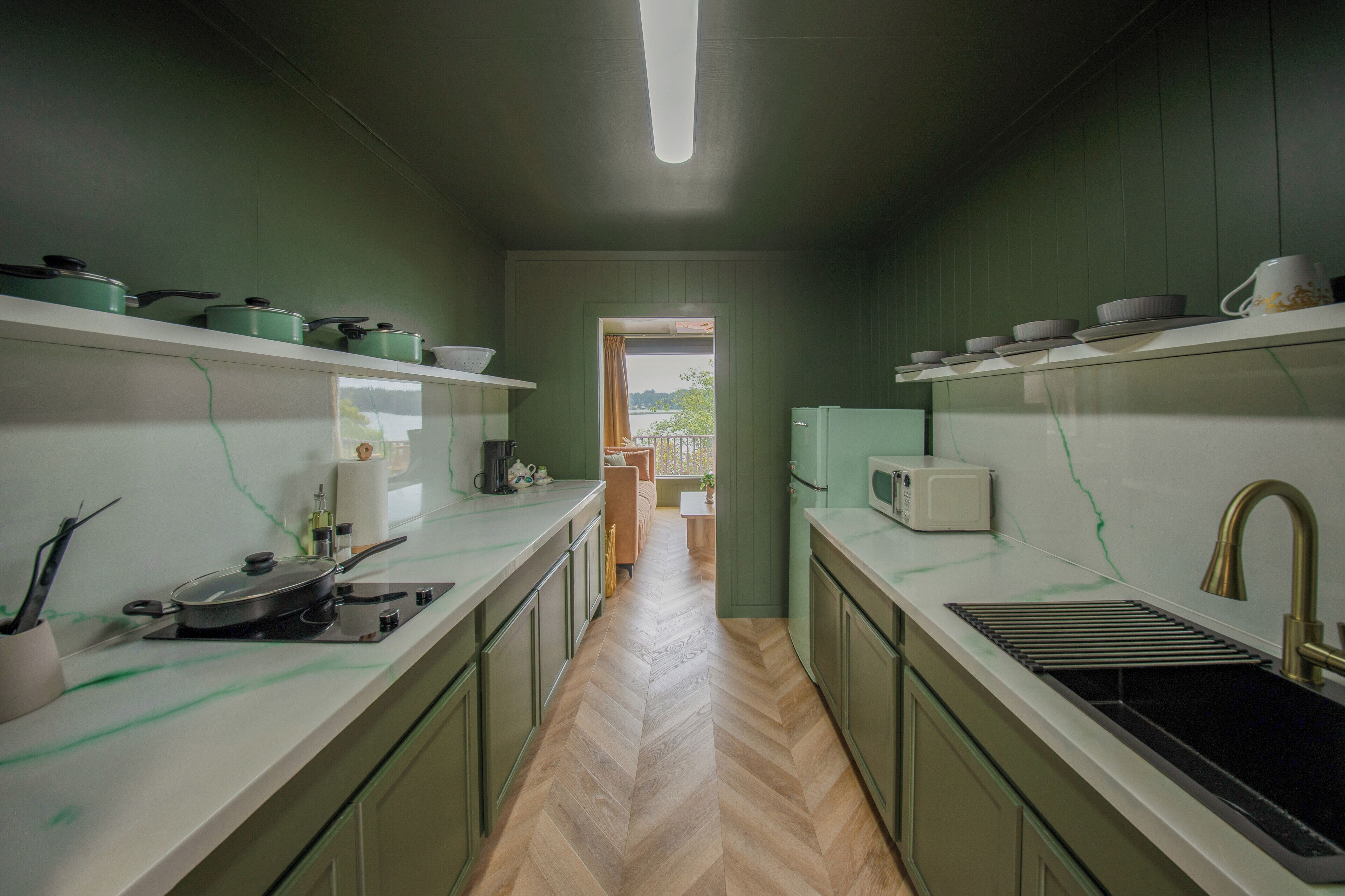 A narrow kitchen with green walls, white countertops, and a herringbone floor. There are shelves with pots and a microwave on the right.