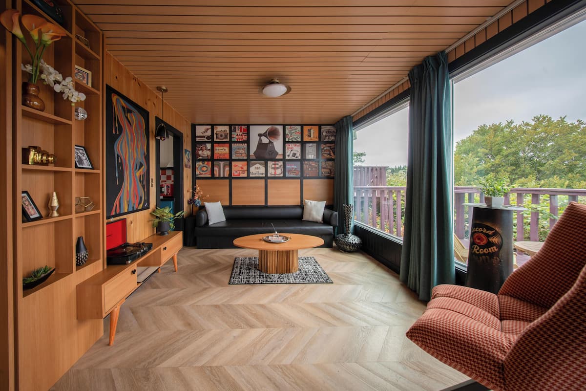 A modern living room with a herringbone wood floor, a round coffee table, a sofa, and a red armchair. There are wooden shelves with decorative items and a large wall mural.