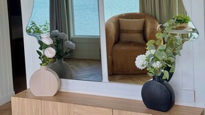 A room with a large arched window offering a view of a lake and forest. A brown sofa is positioned near the window, and there are two vases with flowers on a wooden console table. - The Florentine Waterfront Hotel (Dunes City)