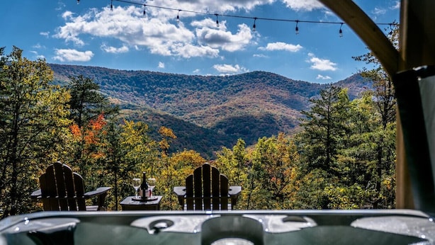 Un bain à remous agrémenté de deux fauteuils Adirondack et d’une table, le tout installé devant un décor de feuillages aux couleurs automnales et d’un paysage montagneux.