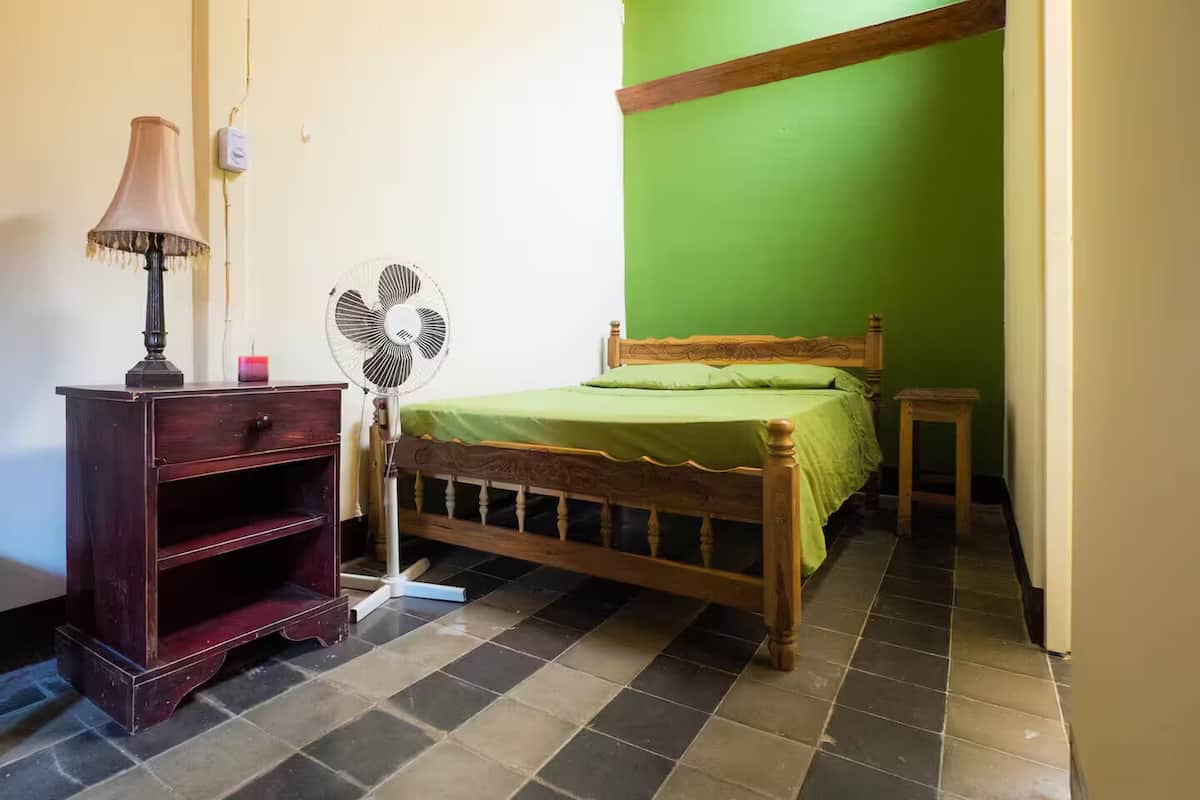 Una habitación con una cama de madera, una mesita de noche, una lámpara y un ventilador de pie.