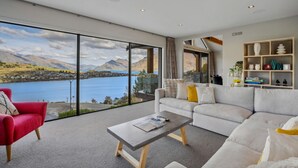 A modern living room with a large sliding glass door leading to a scenic lake view, a red armchair, a grey sofa, and a wooden coffee table.