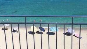 A balcony with a view of the sea, a sandy beach, and several umbrellas.