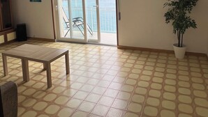 A spacious room with a tiled floor, a ceiling fan, a large sliding glass door leading to a balcony with a view of the sea, a potted plant, and a framed picture on the wall.