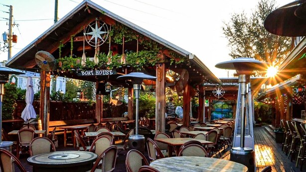 An outdoor seating area with round tables and wicker chairs, a wooden structure labeled "Boat House," and a sign for "At Our House Restaurant." - High Springs Hideaway – 4BR w/ Pool & Bar • Near Springs & UF • Walk to Town (High Springs)