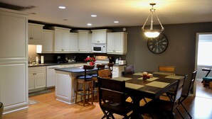 A kitchen with white cabinets, a central island with stools, a dining table with chairs, and a clock on the wall. - High Springs Hideaway – 4BR w/ Pool & Bar • Near Springs & UF • Walk to Town (High Springs)