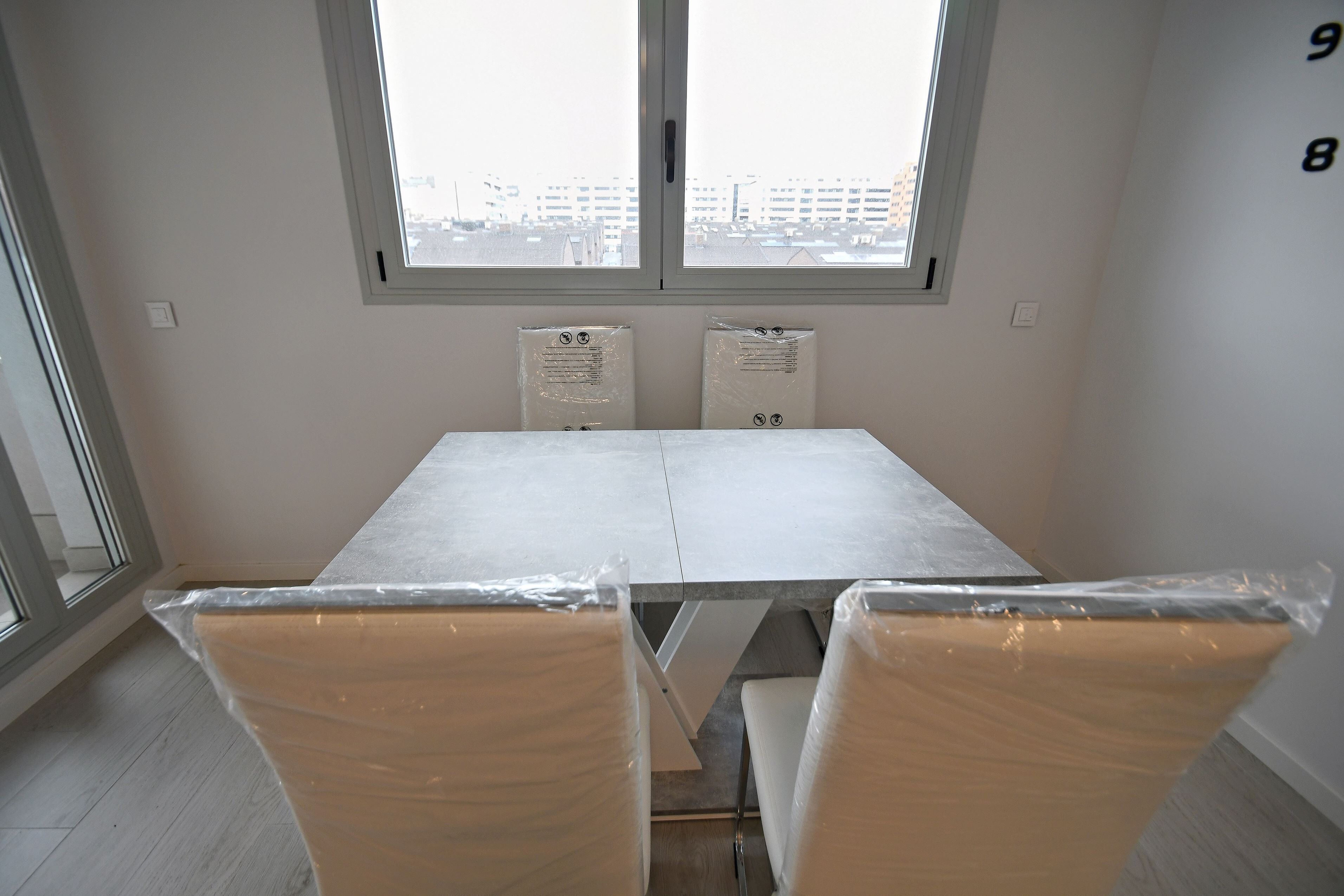Una habitación con un ventanal grande, una mesa blanca y dos sillas cubiertas de plástico.