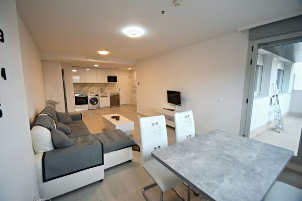 A modern living room with a sofa, a dining table, and a TV. There is a kitchen area with appliances and a laundry machine.
