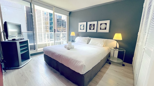 A hotel room with a large bed, a TV on a stand, two bedside lamps, and a window with a view of buildings. - Sydney CBD 2Bedroom Apartment (Sydney)