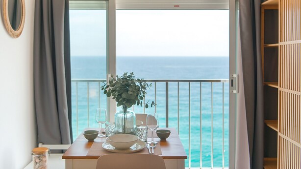 A dining table set with plates, bowls, and glasses, a vase with flowers, and a view of the ocean through a balcony door. - WELCS ESTUDIO 237 PDA junto al mar (Platja d´Aro)