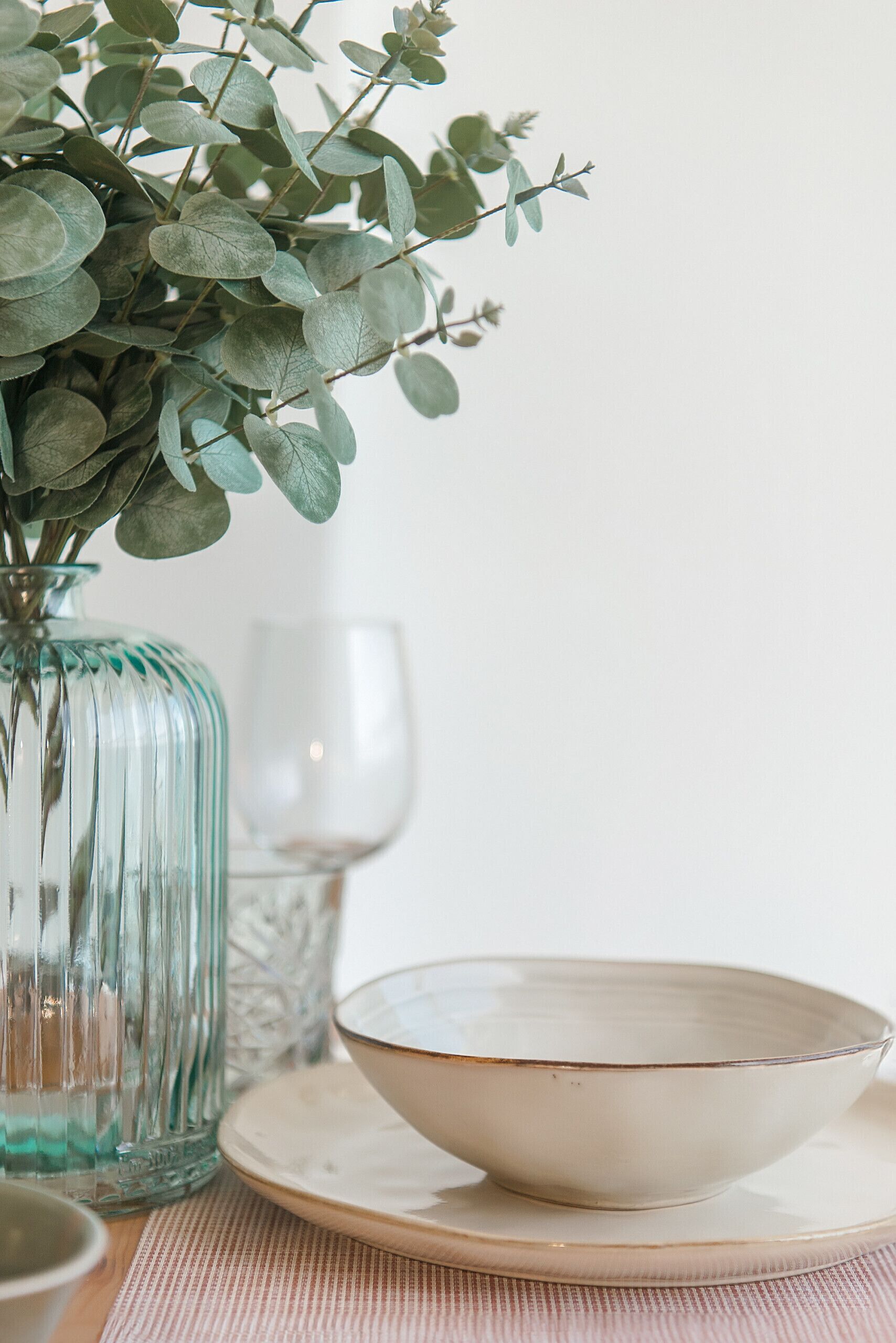 Una mesa dispuesta con un florero de vidrio con hojas verdes, un vaso transparente y un cuenco blanco sobre un plato.