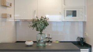 A kitchen with white cabinets, a microwave, a vase with green foliage, and two wine glasses on a dark countertop. - WELCS ESTUDIO 237 PDA junto al mar (Platja d´Aro)