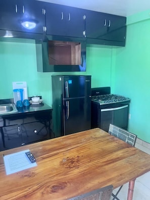 A kitchen with a black refrigerator, stove, and cabinets. There is a wooden table with a remote control and a menu on it. - Garden Lodge Hotel (Placencia)