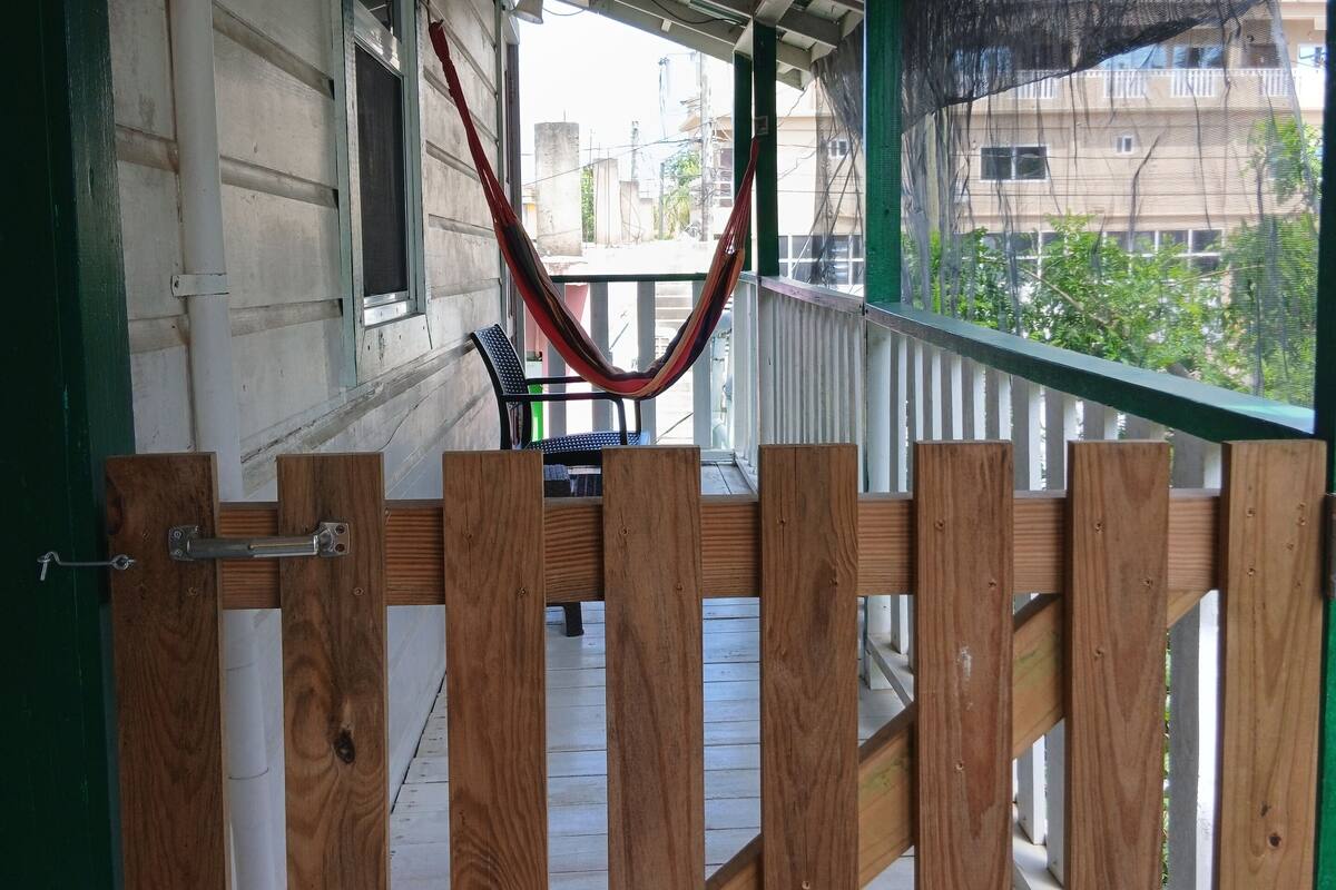 Un porche de madera con una hamaca, una entrada con portón y vistas a edificios al fondo.