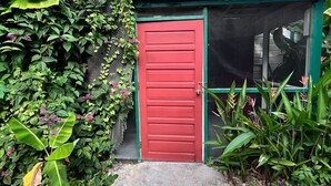 Una puerta roja con techo verde, rodeada de follaje verde exuberante y flores rosadas.