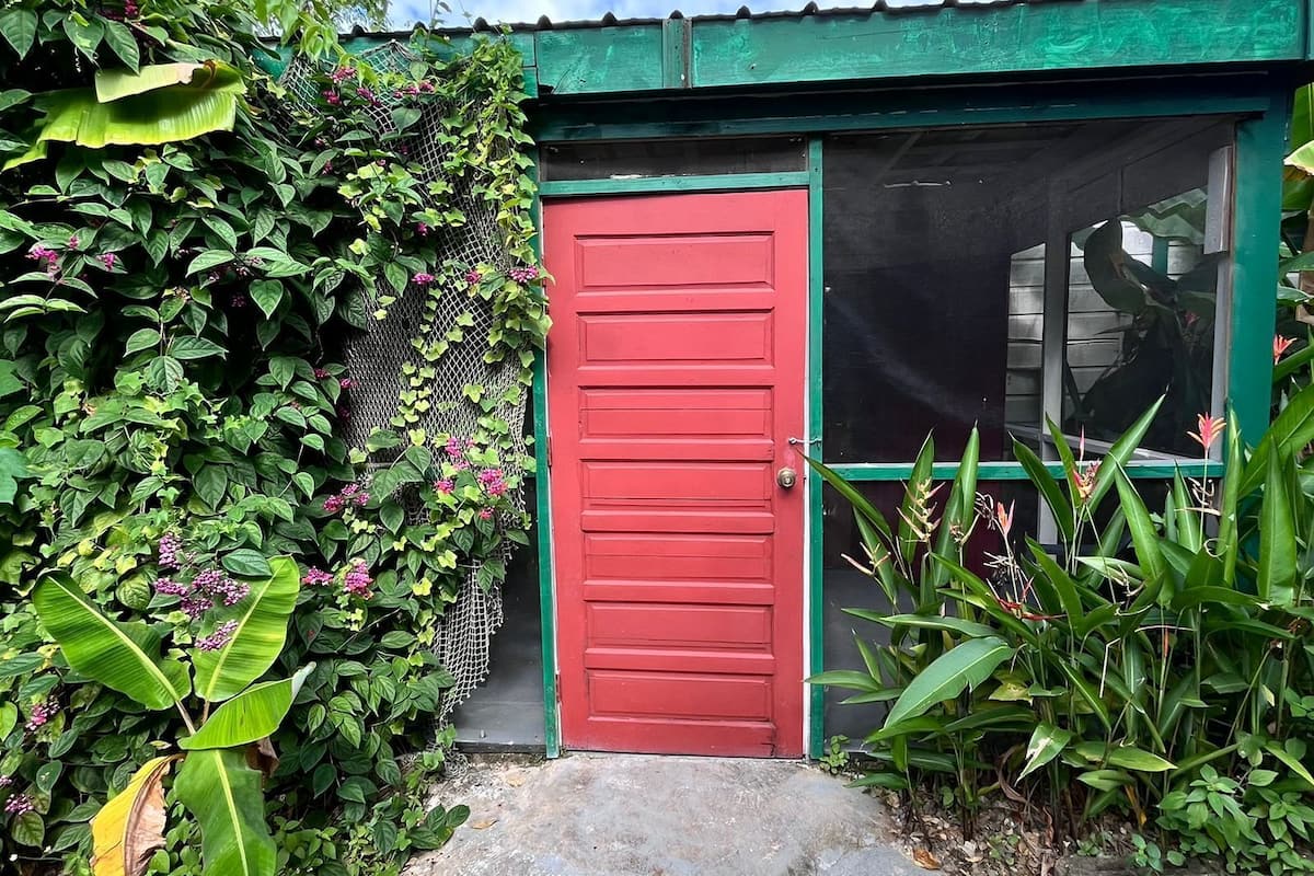 A red door with a green roof, surrounded by lush green foliage and pink flowers.