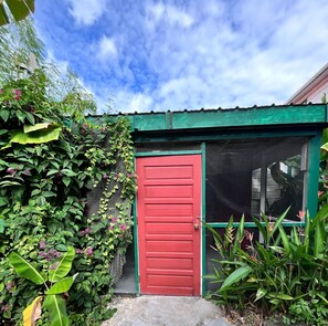 Una puerta roja con techo verde, rodeada de follaje verde exuberante y flores rosadas.