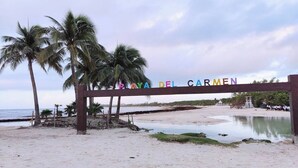 Ingresso sulla spiaggia, con palme e un cartello che riporta "PAYA DEL CARMEN".