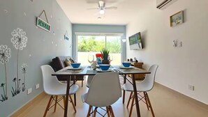 Zona pranzo con tavolo apparecchiato per quattro persone, sedie bianche, ventilatore a soffitto e una grande finestra vista sul verde.