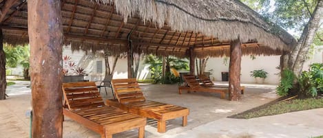 A covered outdoor seating area with wooden lounge chairs and a thatched roof structure.