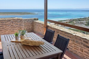 Ein Holztisch mit Stühlen, ein Blumentopf und Weingläser auf einem Balkon mit Blick aufs Meer.
