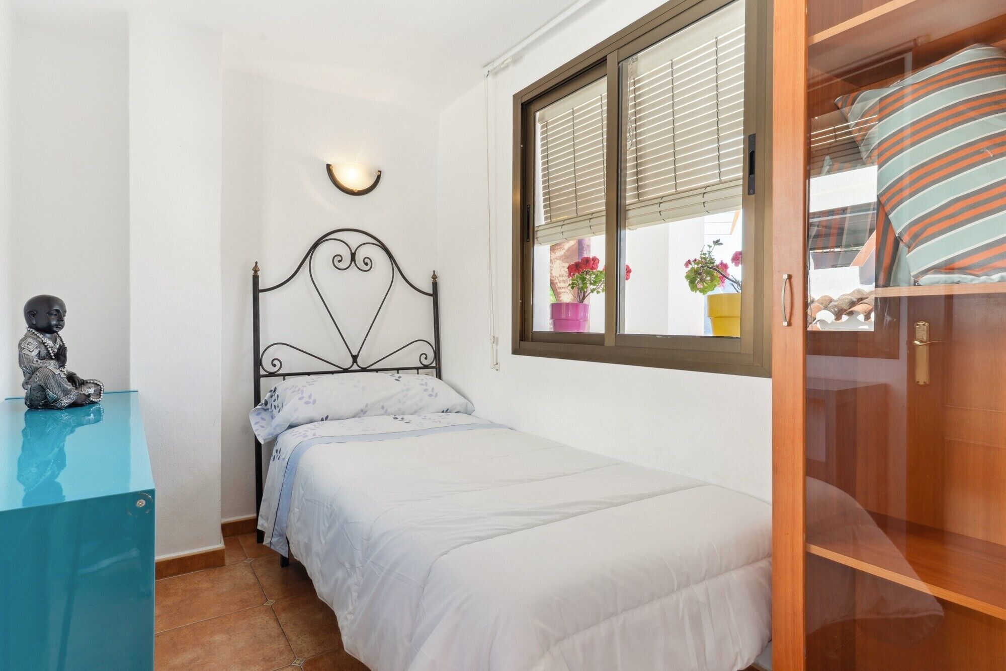 A bedroom with a single bed, a nightstand, a wall-mounted light, a window with blinds, and a view of the outside.