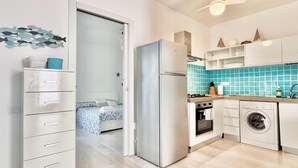 Una cocina moderna con refrigerador, horno y lavadora, un dormitorio con cama y mesita de noche, y un pasillo con puerta que da al dormitorio.