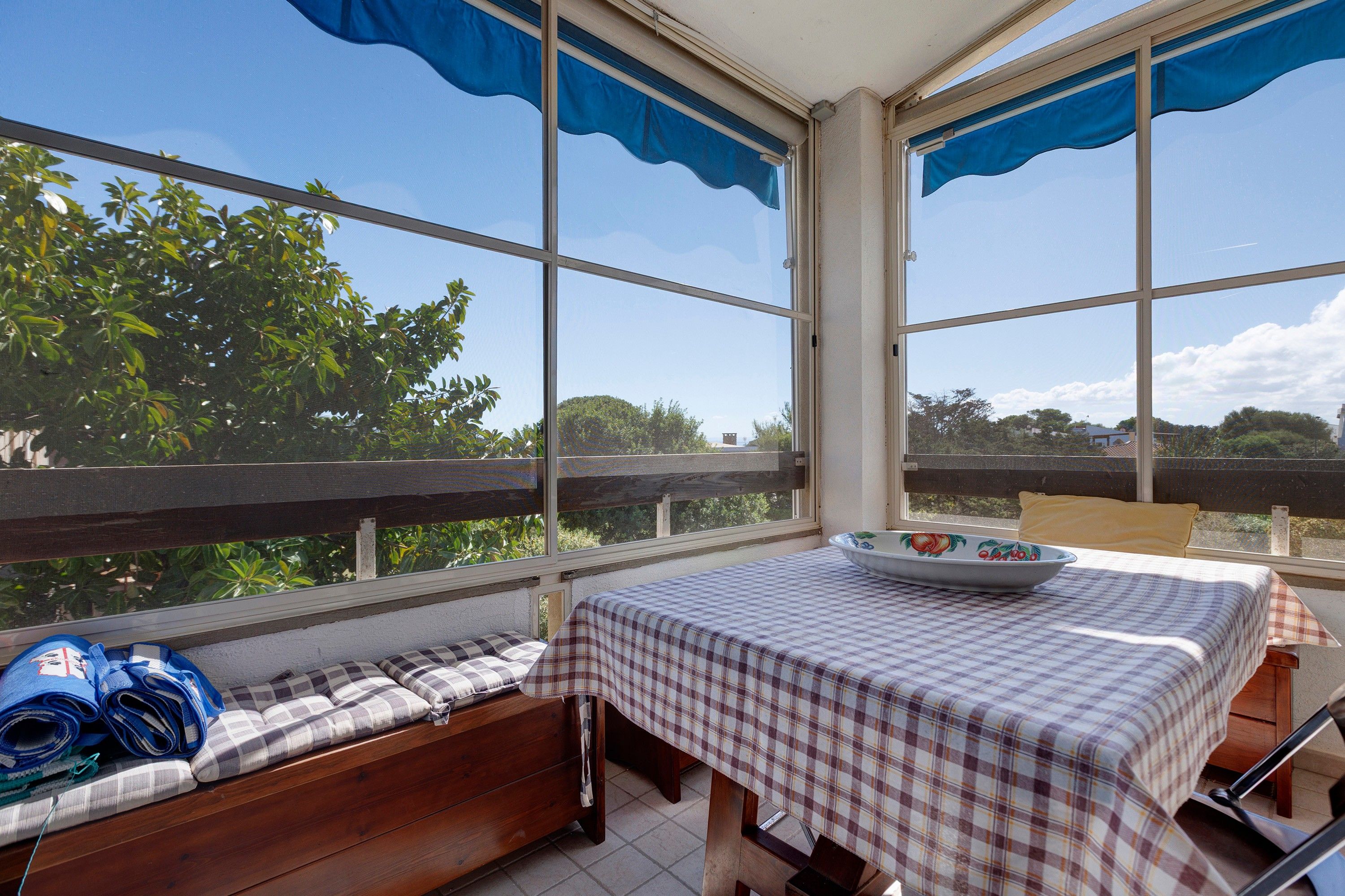Una habitación iluminada por el sol, con un banco, una mesa con mantel a cuadros y vistas al exterior con vegetación.