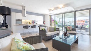 Un moderno espacio de sala con un sofá, sillas, una mesa de centro de vidrio y una cocina al fondo.