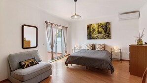 A bedroom with a bed, a sofa, a window with curtains, a painting, a mirror, a lamp, and an air conditioner.