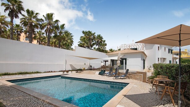 A swimming pool with a tiled deck, lounge chairs, and a covered area with a table and chairs.