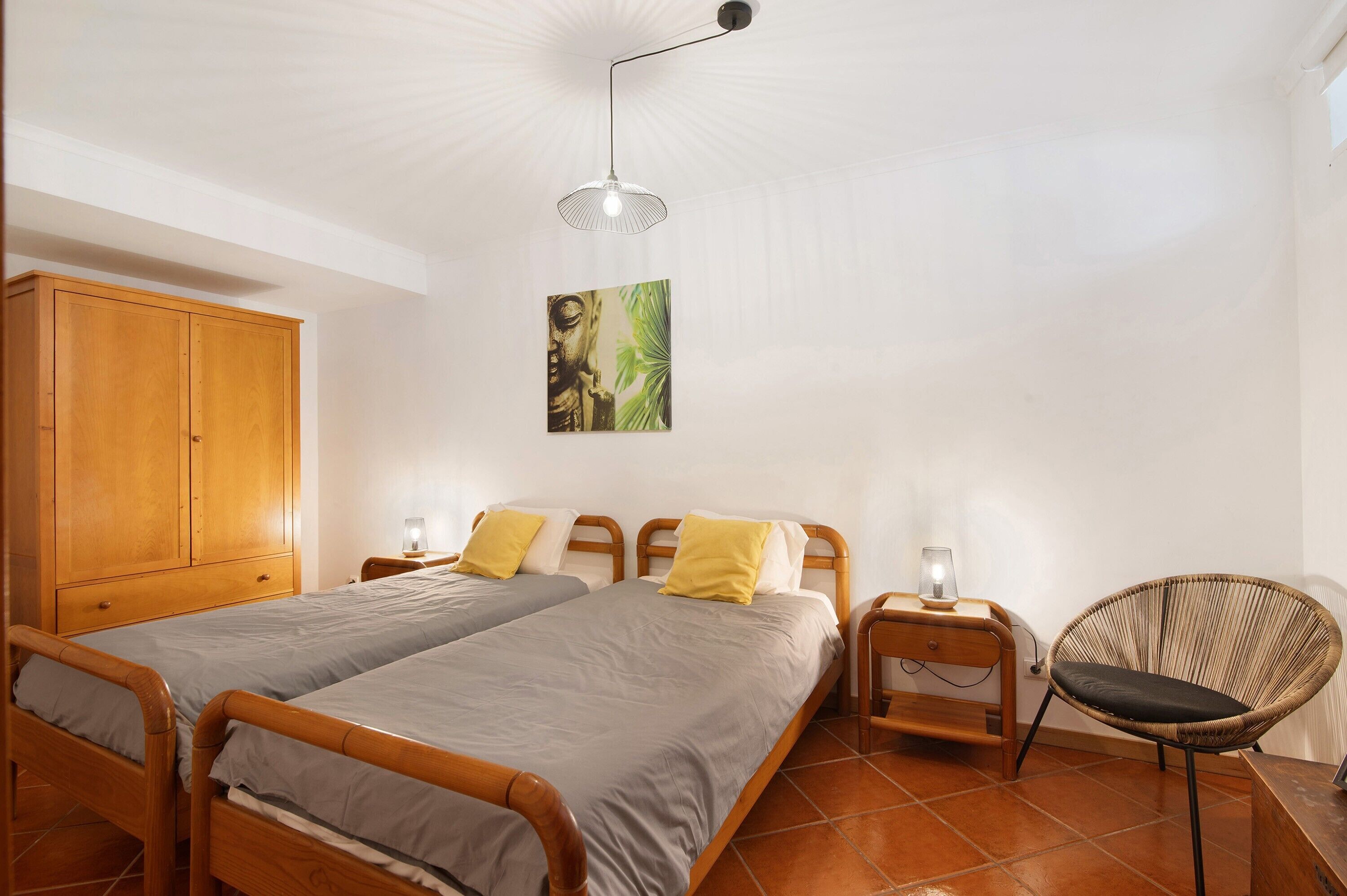 Una habitación con dos camas de madera, un armario de madera, una silla de ratán, un cuadro colgado en la pared y una lámpara del techo.