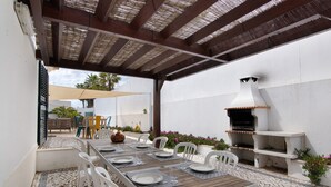An outdoor patio with a long wooden table set for dining, surrounded by white chairs, a barbecue grill, and a white wall.