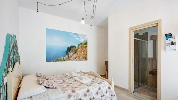 Ein Schlafzimmer mit einem Bett, einem Gemälde einer Küstenlandschaft, einer Holztür und einem kleinen Regal mit einer Fernbedienung.