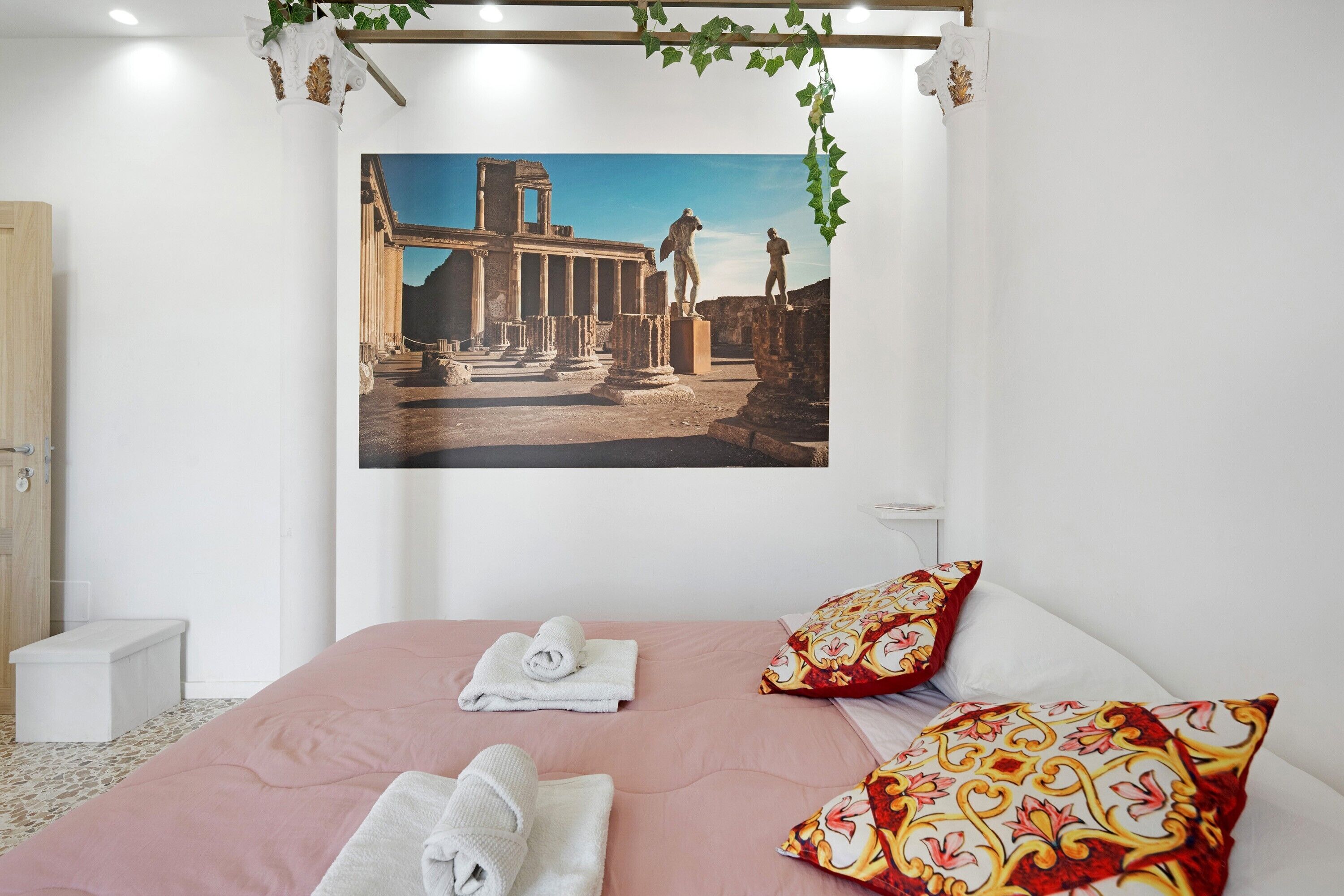 A bedroom with a bed, pillows, a wall-mounted picture of ancient ruins, a wooden door, and a small white bench.