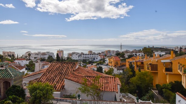 A coastal cityscape with terracotta-roofed buildings, a clear sky, and a view of the sea.