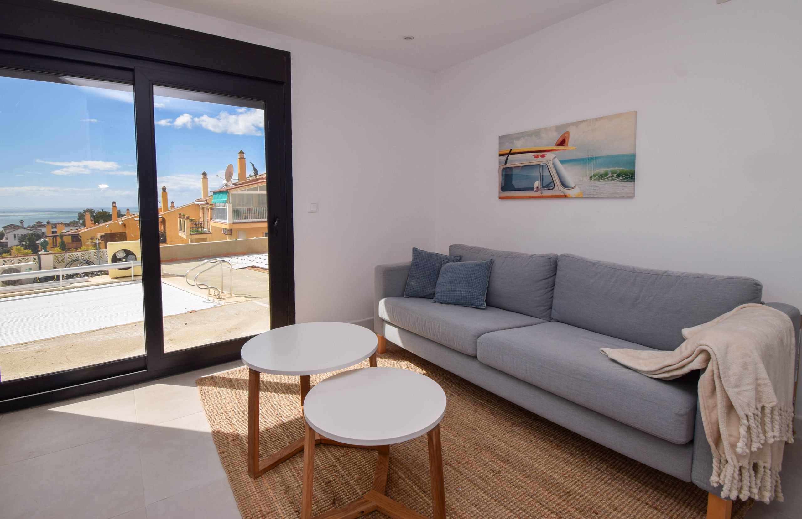A modern living room with a grey sofa, two white round tables, and a large window offering a view of buildings and a clear sky.