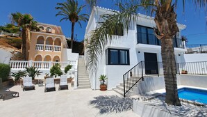 A modern two-story building with a pool, surrounded by palm trees and potted plants.