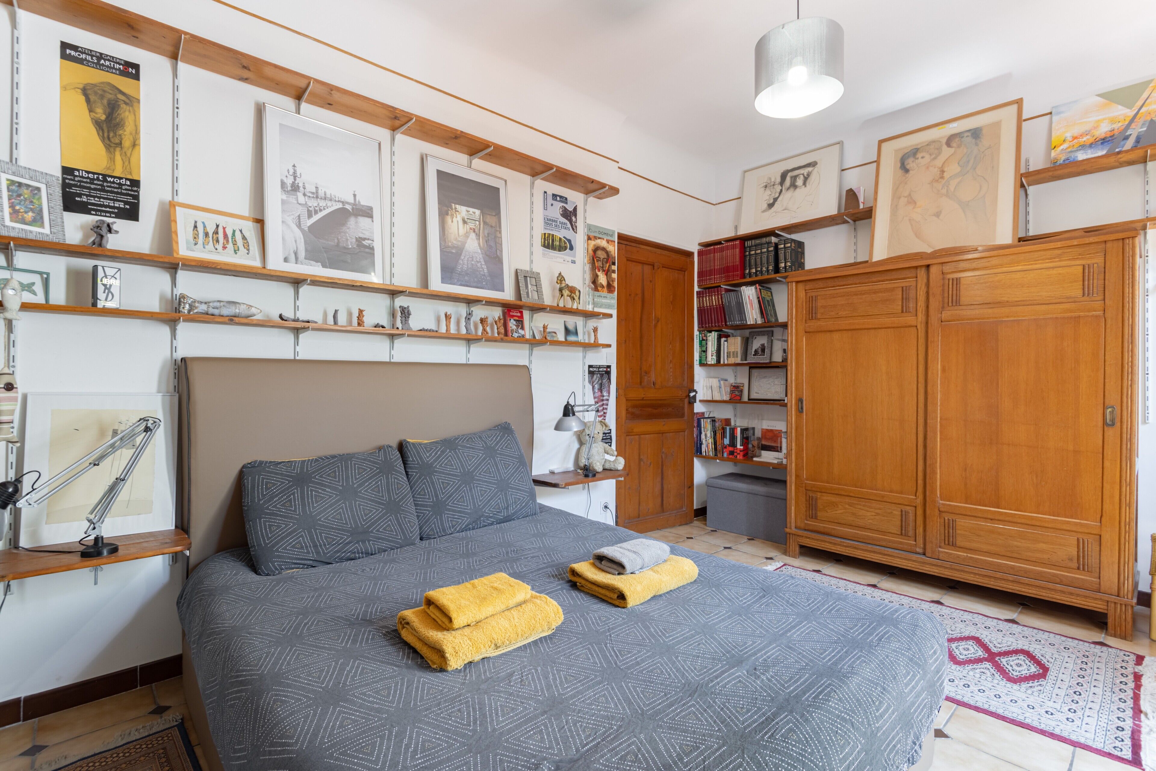 A bedroom with a bed, a wooden wardrobe, shelves with books and pictures, and a desk with a lamp.