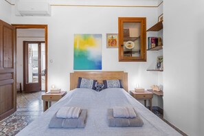 Une chambre à coucher avec un grand lit, une tête de lit en bois, une table de chevet, une commode, une étagère fixée au mur et une porte donnant sur une autre pièce.
