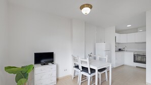 Une cuisine moderne avec des meubles blancs, une table à manger avec des chaises, un téléviseur posé sur une commode et une plante en pot.