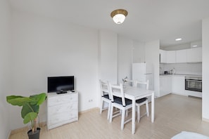A modern kitchen with white cabinets, a dining table with chairs, a TV on a dresser, and a potted plant.