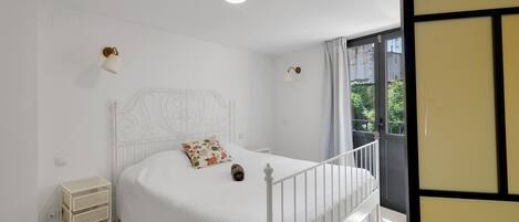 Una cama blanca con cabecera, una mesita de noche pequeña, dos lámparas fijadas a la pared y vistas de vegetación al exterior.