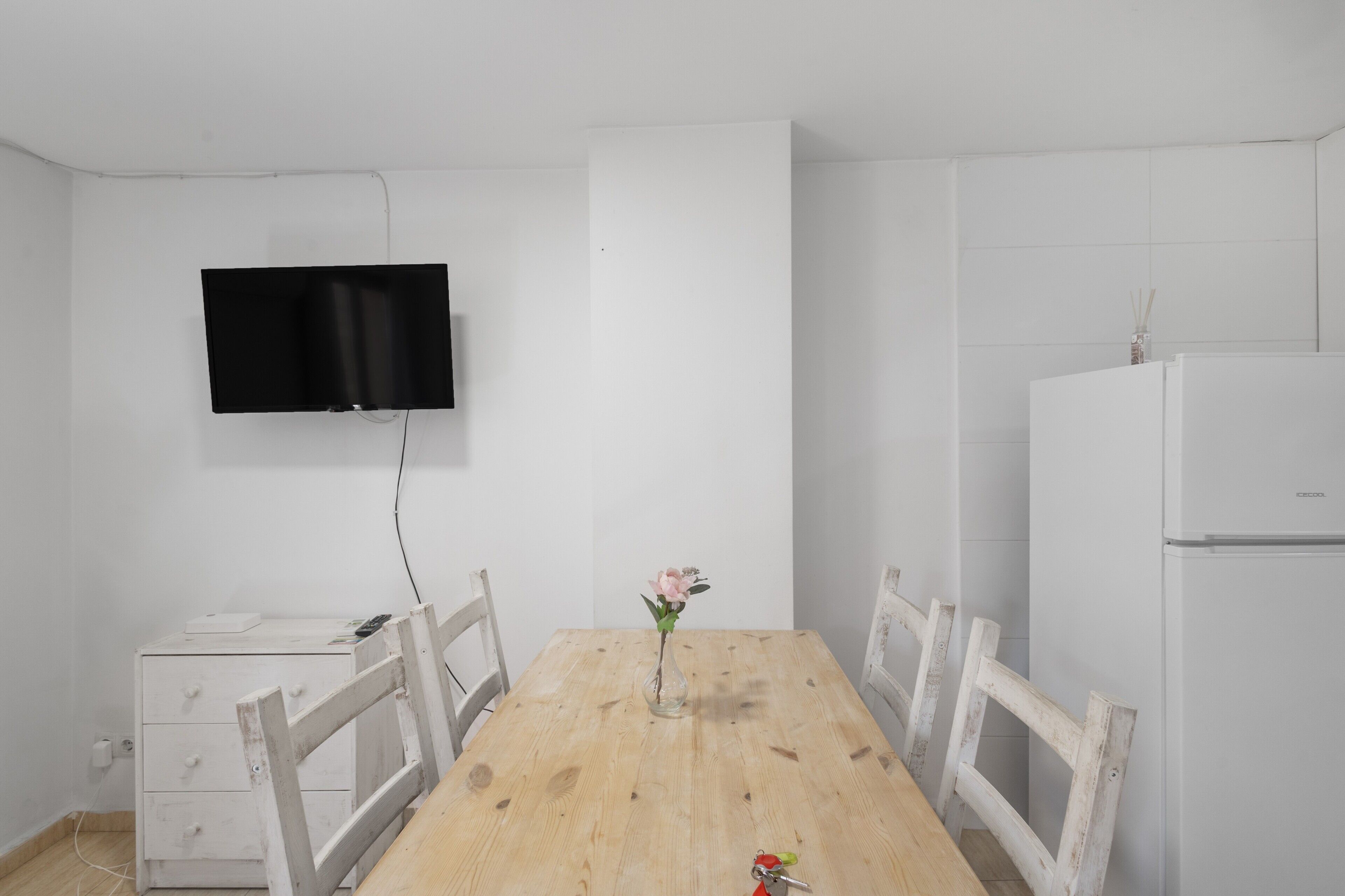 Een eethoek met een houten tafel, stoelen, een aan de muur bevestigde televisie en een witte koelkast.