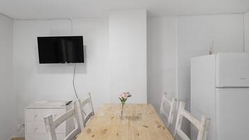 Een eethoek met een houten tafel, stoelen, een aan de muur bevestigde televisie en een witte koelkast.